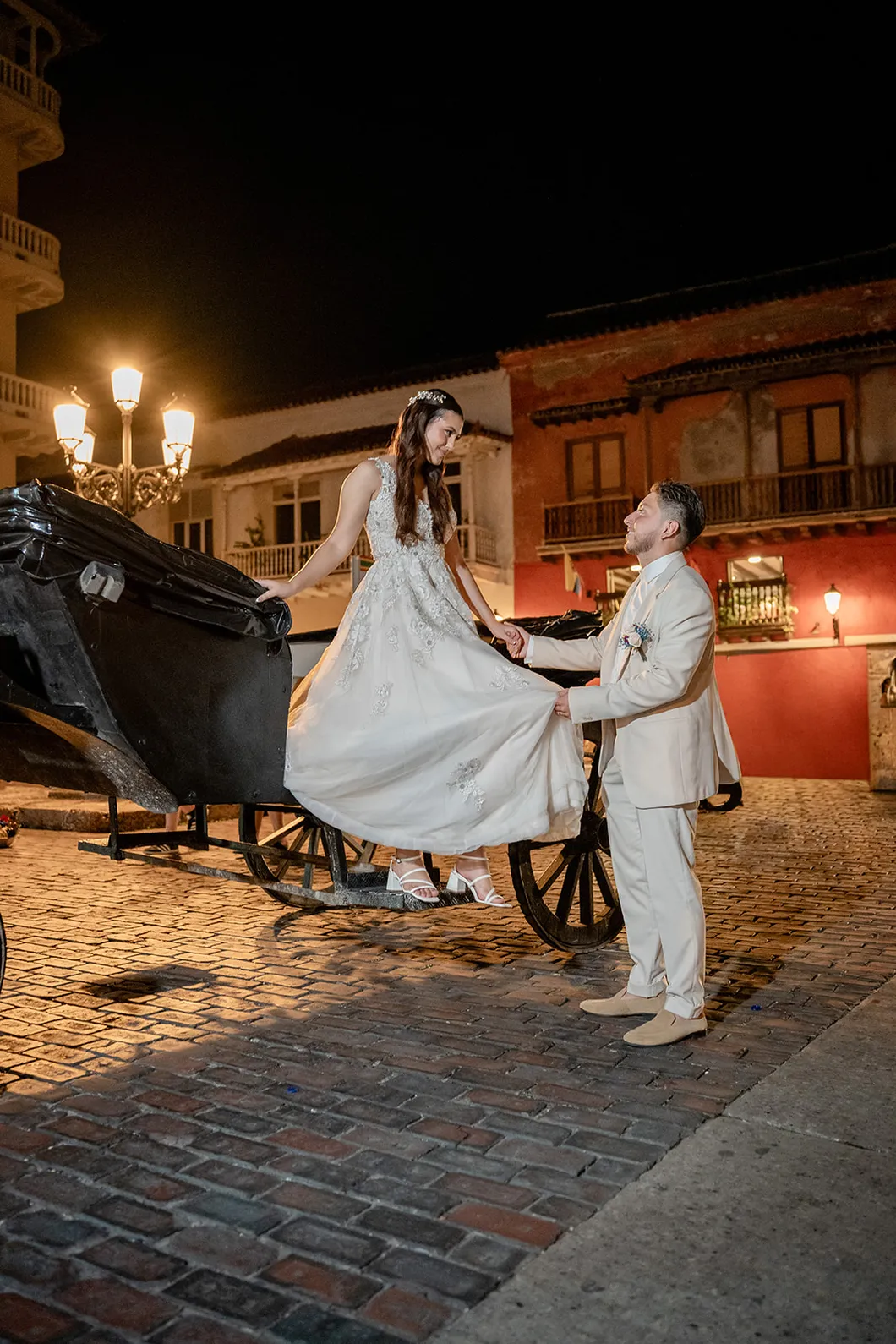 La novia baja del carruaje mientras el novio le sostiene el vestido, de noche en Cartagena, por Omar Fraija